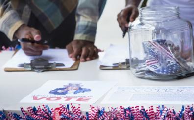 Two Black people stand behind a white table. They are both checking through clipboards with a pen. There is a glass jar in front of them with little American flags in it, and little flags line the front edge of the table as well.