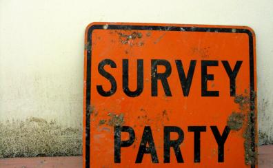 A dirty, beat-up orange street sign leans againts a beige wall. Black letters on the sign read "SURVEY PARTY."
