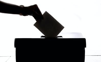 A backlit image of a hand dropping a piece of paper into a ballot box.