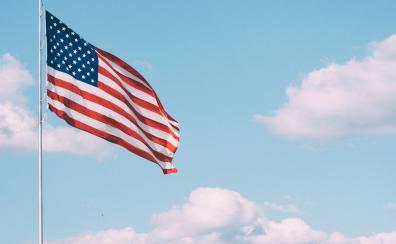 A U.S. flag waves gently on a tall silver pole. The sky behind it is blue, with a few fluffy white clouds.