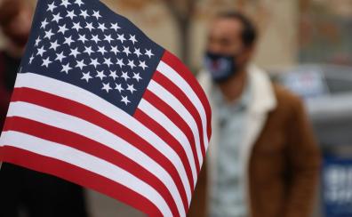 A handheld American flag is held in front of the camera. Slightly blurred in the background, we can see a man in a brown jacket with a white collar who is wearing a mask over his nose and mouth.