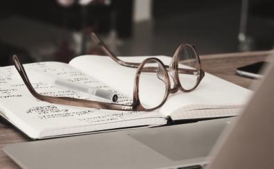 A pair of glasses lies upside down on top of a lined notebook. The notebook is open and one page is covered in writing. A pen rests next to the glasses.