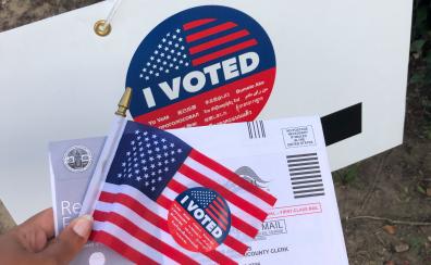 Hand holding an American flag and a mail ballot return envelope. 