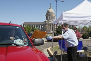 A masked and gloved man takes an envelope from a man in a car in front of a capitol building