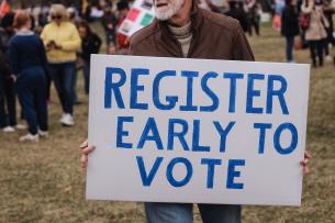 A man holds a hand-painted sign that reads "Register early to vote"