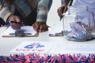 Two Black people stand behind a white table. They are both checking through clipboards with a pen. There is a glass jar in front of them with little American flags in it, and little flags line the front edge of the table as well.