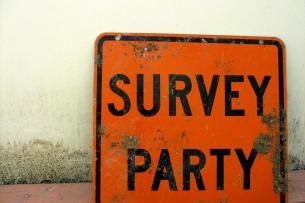 A dirty, beat-up orange street sign leans againts a beige wall. Black letters on the sign read "SURVEY PARTY."