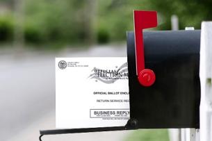 A black mailbox with its red arm raised up. The mailbox is open, and an absentee ballot is halfway in, as though it's just been slipped into the box.