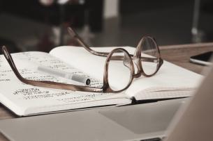 A pair of eyeglasses with thick brown translucent frames rests, upside down, on an open notebook, where one of the pages is full of scribbled notes.