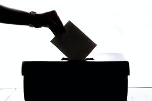 A backlit image of a hand dropping a piece of paper into a ballot box.