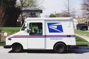 A white USPS truck is stopped at the curb in a suburban neighborhood.