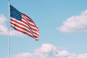 A U.S. flag waves gently on a tall silver pole. The sky behind it is blue, with a few fluffy white clouds.