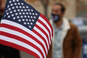 A handheld American flag is held in front of the camera. Slightly blurred in the background, we can see a man in a brown jacket with a white collar who is wearing a mask over his nose and mouth.