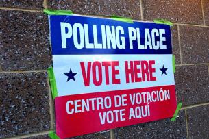 A brick wall with a sign taped to it with green masking tape. The sign is striped red, white, and blue; the text on the sign reads "Polling place, vote here. Centro de votacion, vote aqui."