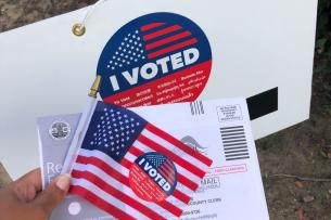 Hand holding an American flag and a mail ballot return envelope. 